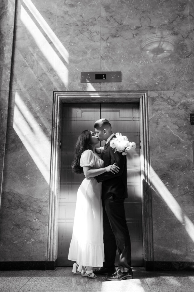 a black and white photo of a male and female kissing in front of the golden doors at the San Diego Courthouse. 